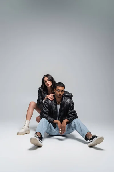 Full length of stylish woman in autumnal outfit sitting with young african american man on grey — Stock Photo