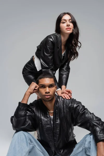 Mujer con estilo en chaqueta de cuero posando con el hombre joven afroamericano aislado en gris - foto de stock