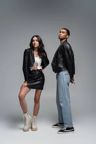 Full length of stylish woman in leather outfit and boots posing with young african american man on grey — Stock Photo