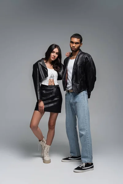 Pleine longueur de jeune femme en tenue en cuir et bottes posant avec l'homme afro-américain élégant sur gris — Photo de stock