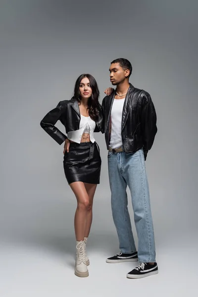 Full length of young woman in leather outfit and boots posing with stylish african american model on grey — Stock Photo