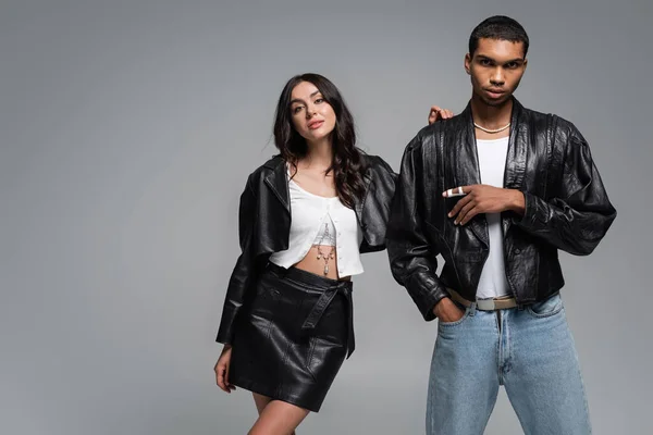 Stylish african american man posing with hand in pocket near young woman in leather outfit isolated on grey — Stock Photo