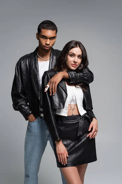 Stylish african american man posing with hand in pocket and hugging young woman in leather outfit isolated on grey — Stock Photo