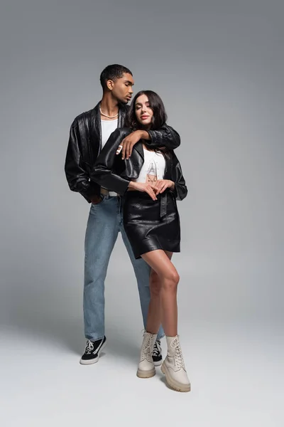 Full length of stylish african american man posing with hand in pocket and hugging young woman in leather outfit on grey — Stock Photo