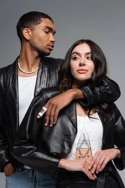 Stylish african american man posing with hand in pocket and hugging brunette woman in leather jacket isolated on grey — Stock Photo