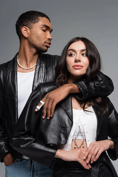 Hombre afroamericano elegante posando con la mano en el bolsillo y abrazando a la mujer morena en chaqueta de cuero aislado en gris - foto de stock