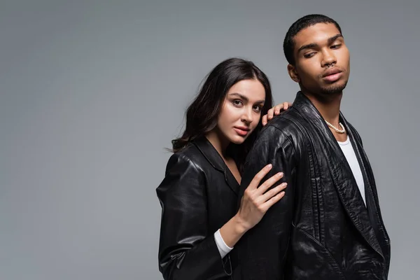 Mujer joven en chaqueta de cuero abrazando elegante hombre afroamericano aislado en gris - foto de stock