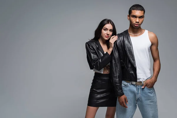 Jeune femme en veste en cuir étreignant élégant homme afro-américain posant avec la main dans la poche de jeans isolés sur gris — Photo de stock