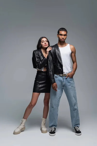 Longitud completa de la mujer en traje de cuero abrazando elegante hombre afroamericano posando con la mano en el bolsillo en gris - foto de stock