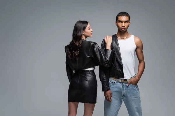 Mujer joven en traje de cuero abrazando elegante hombre afroamericano posando con la mano en el bolsillo aislado en gris - foto de stock