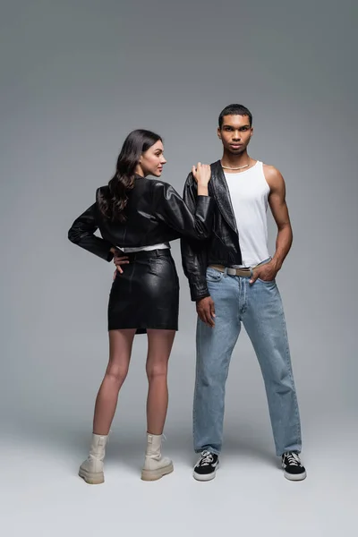 Longitud completa de la mujer en falda de cuero tocando hombro de hombre afroamericano elegante posando con la mano en el bolsillo en gris - foto de stock