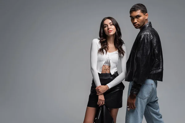 Morena mujer en falda de cuero sosteniendo chaqueta negra cerca de hombre afroamericano elegante aislado en gris - foto de stock