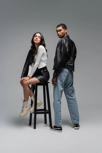 Full length of woman in trendy outfit sitting on high chair near stylish african american man on grey — Stock Photo