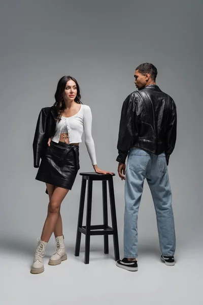 Повна довжина жінки в модному вбранні, що стоїть біля високого стільця і стильний афроамериканський чоловік на сірому — стокове фото