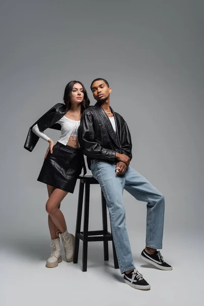 Piena lunghezza della donna in abito di pelle e l'uomo afro-americano elegante in posa vicino seggiolone su grigio — Foto stock