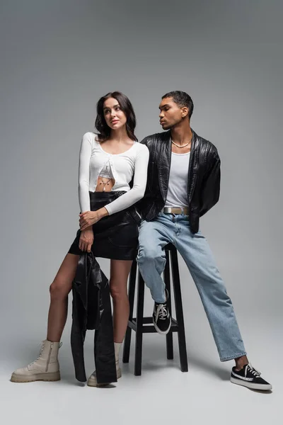 Full length of woman in stylish outfit and young african american man in leather jacket posing near high chair on grey — Stock Photo