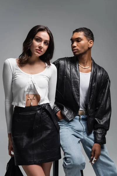 Young woman in stylish outfit and young african american man in leather jacket posing near high chair isolated on grey — Stock Photo