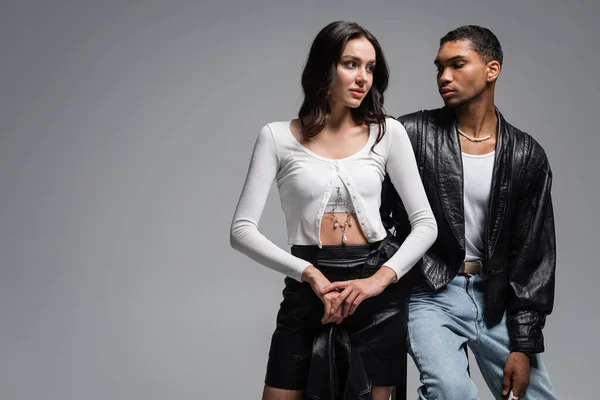 Young woman in stylish outfit near young african american man in leather jacket sitting on high chair isolated on grey — Stock Photo