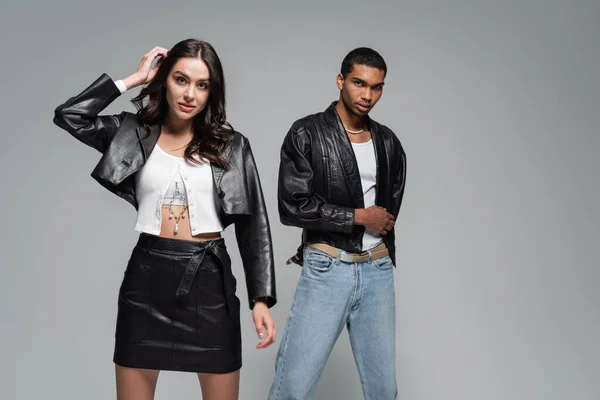 Young and stylish multiethnic couple in leather jackets posing isolated on grey — Stock Photo