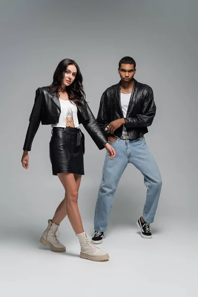 Full length of young and stylish multiethnic couple in leather jackets posing on grey — Stock Photo