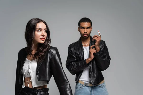 Young and stylish multiethnic couple in black leather jackets posing isolated on grey — Stock Photo