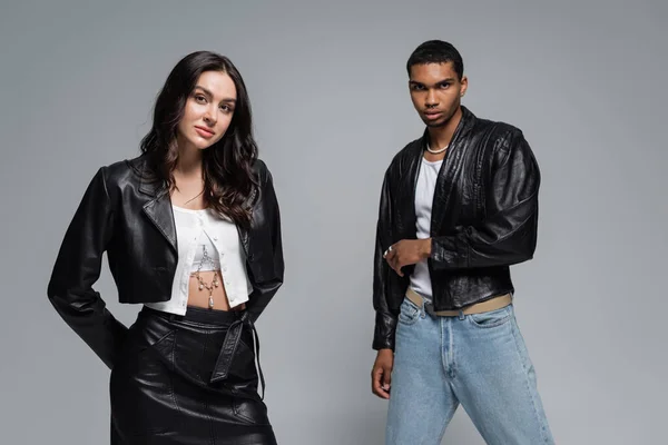 Young multiethnic couple in black leather jackets looking at camera isolated on grey — Stock Photo