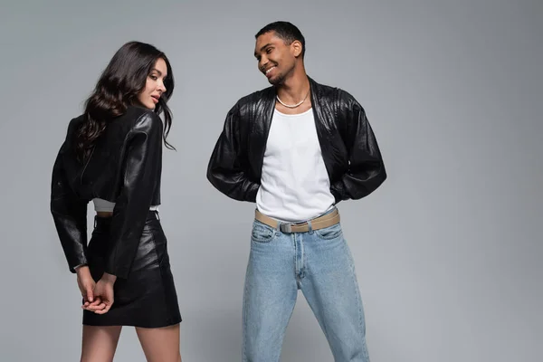 Hombre afroamericano feliz mirando a mujer joven con estilo en traje de cuero aislado en gris - foto de stock