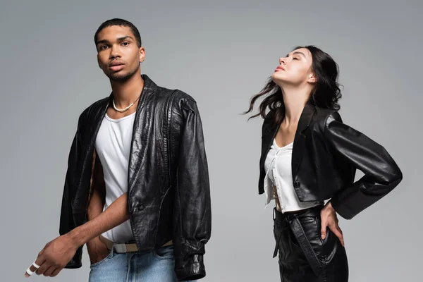 Bonita joven posando con los ojos cerrados cerca de hombre afroamericano en chaqueta de cuero aislado en gris - foto de stock