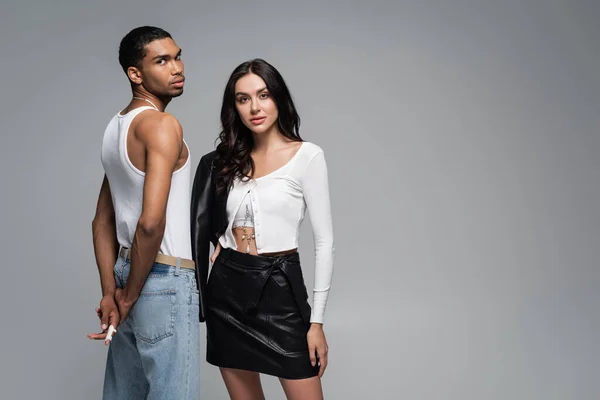 Young woman in stylish outfit posing near african american man in tank top and jeans isolated on grey — Stock Photo