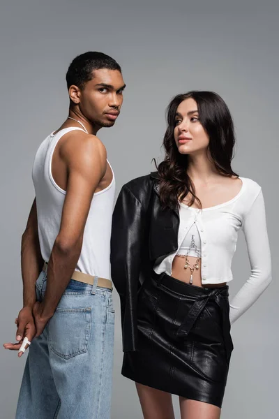 Jovem mulher na roupa da moda posando perto de homem americano africano em top tanque e jeans isolados em cinza — Fotografia de Stock