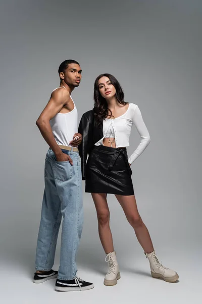 Full length of young woman in stylish outfit posing near african american man in tank top and jeans on grey — Stock Photo