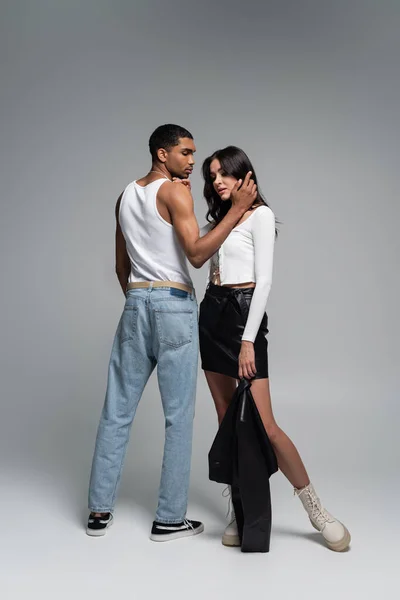 Pleine longueur de jeune homme afro-américain en débardeur étreignant femme en tenue élégante sur gris — Photo de stock