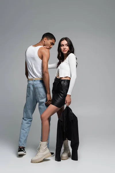 Full length of brunette woman in stylish outfit hugging young african american man in tank top on grey — Stock Photo