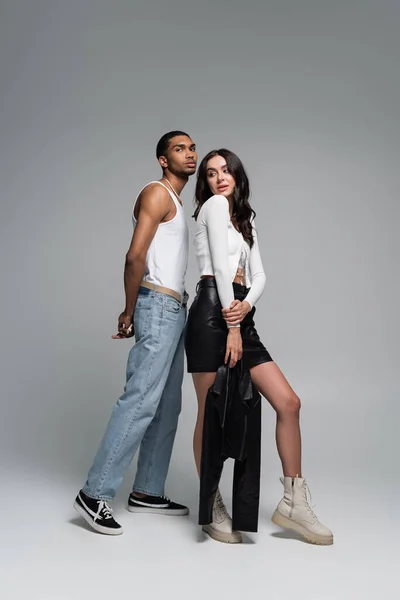 Longitud completa de joven afroamericano hombre en camiseta sin mangas y mujer morena en traje elegante posando en gris - foto de stock