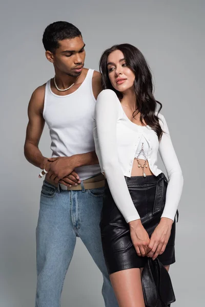 Young african american man in tank top looking at pretty woman in leather skirt  isolated on grey — Stock Photo