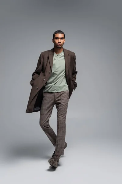 Comprimento total do jovem afro-americano em casaco de lã outonal olhando para a câmera e posando com as mãos nos bolsos em cinza — Fotografia de Stock