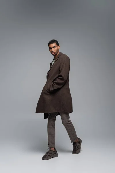 Pleine longueur de jeune homme afro-américain en manteau de laine automnal regardant la caméra et posant la main dans la poche sur le gris — Photo de stock