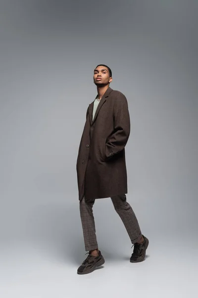 Pleine longueur de jeune homme afro-américain en manteau d'automne regardant la caméra et posant la main dans la poche sur le gris — Photo de stock