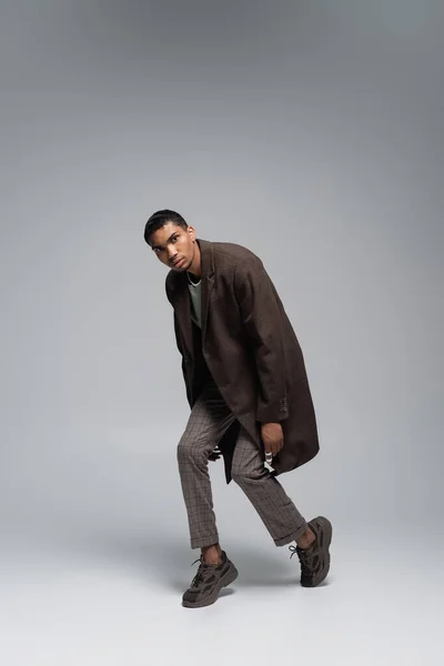 In voller Länge junger afrikanisch-amerikanischer Mann in herbstlichem Wollmantel, der in die Kamera schaut und auf grau posiert — Stockfoto