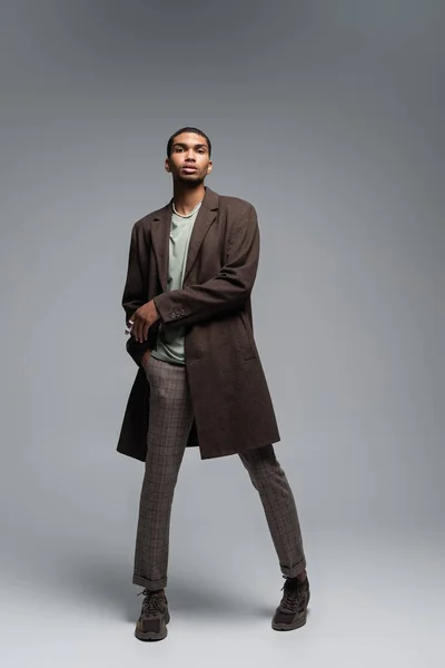 Pleine longueur de jeune homme afro-américain en manteau de laine automnal regardant la caméra et debout avec la main dans la poche sur le gris — Photo de stock