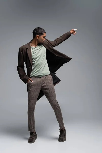 Full length of young african american man in autumnal woolen coat posing with hand in pocket on grey — Stock Photo