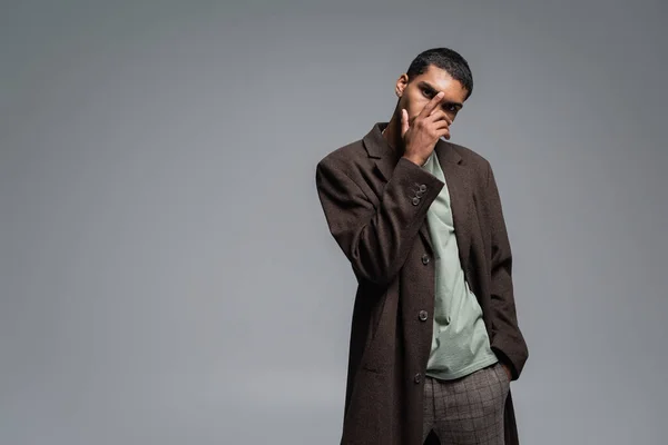 Young african american man in woolen coat looking at camera while posing with hand in pocket isolated on grey — Stock Photo