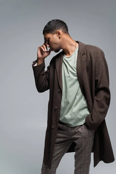 Young african american man in coat touching face while posing with hand in pocket isolated on grey — Stock Photo