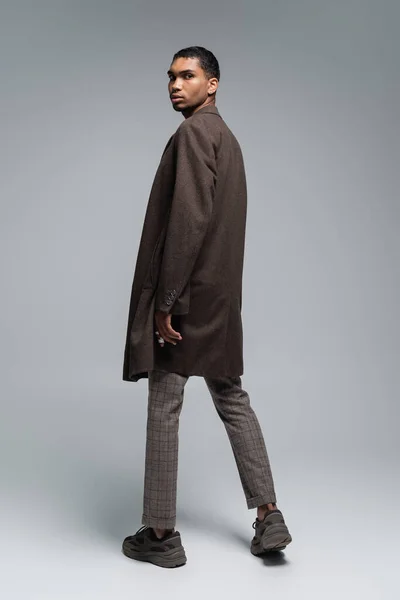 Повна довжина стильного афроамериканця в автентичному вовняному пальто дивиться на камеру і стоїть на сірому — стокове фото