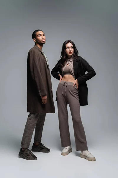 Longitud completa de elegantes modelos interracial en trajes otoñales posando juntos en gris - foto de stock