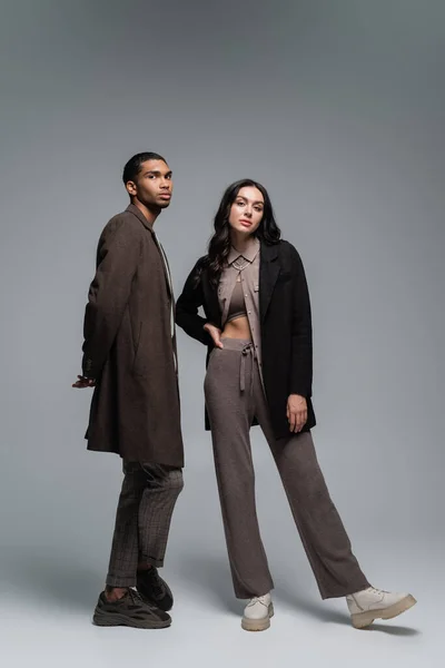 Longitud completa de elegantes modelos multiétnicos en trajes otoñales posando juntos en gris - foto de stock