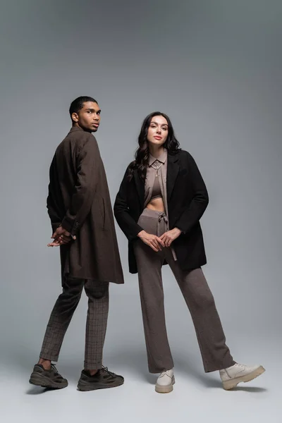 Longitud completa de elegantes modelos interracial en trajes otoñales de moda posando juntos en gris - foto de stock