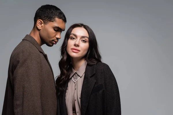 Joven africano americano hombre en abrigo cerca elegante mujer en chaqueta aislado en gris - foto de stock