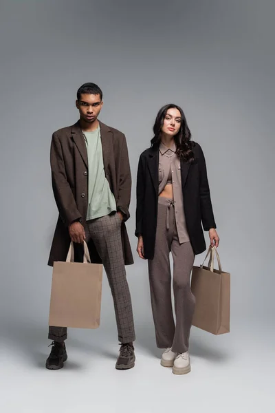 Full length of stylish interracial man and woman in autumnal outfits holding shopping bags on grey — Stock Photo