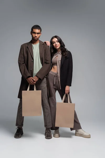 Full length of stylish interracial couple in trendy outfits holding shopping bags on grey — Stock Photo